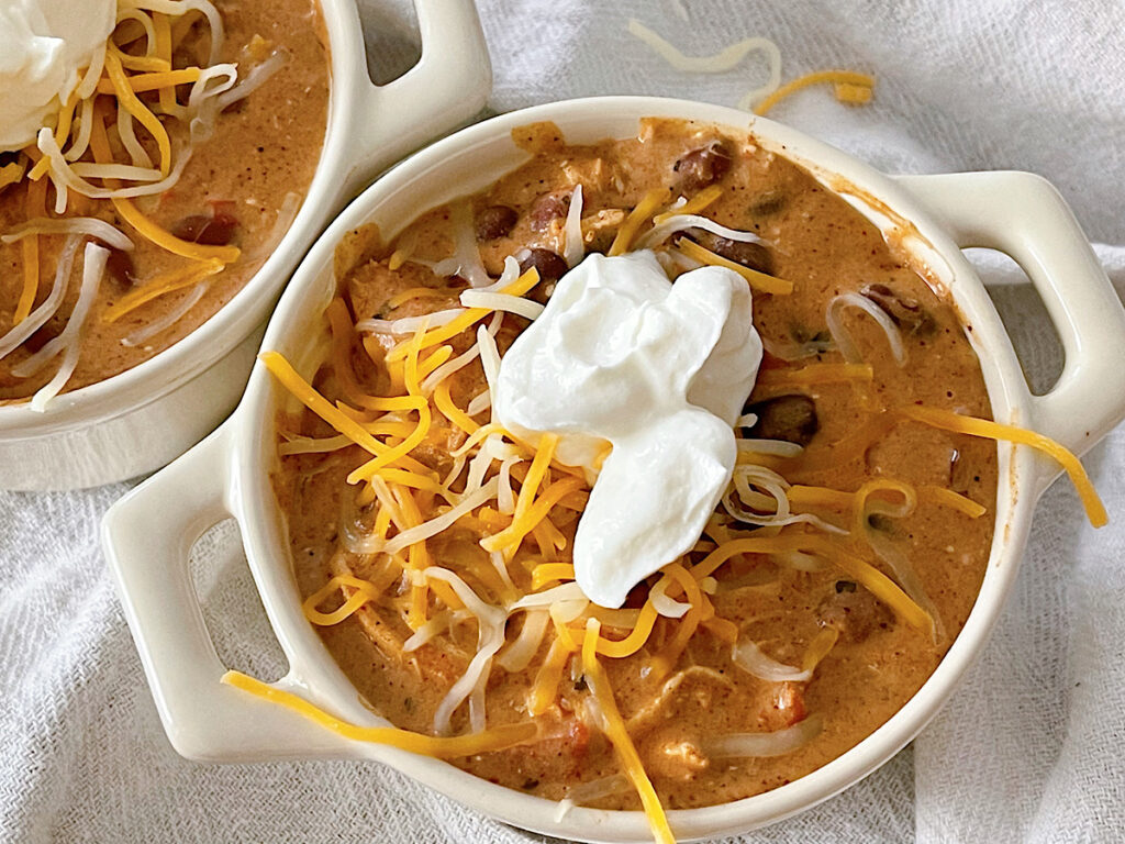 Two bowls of Cream Cheese Chicken Chili topped with shredded cheese and sour cream.