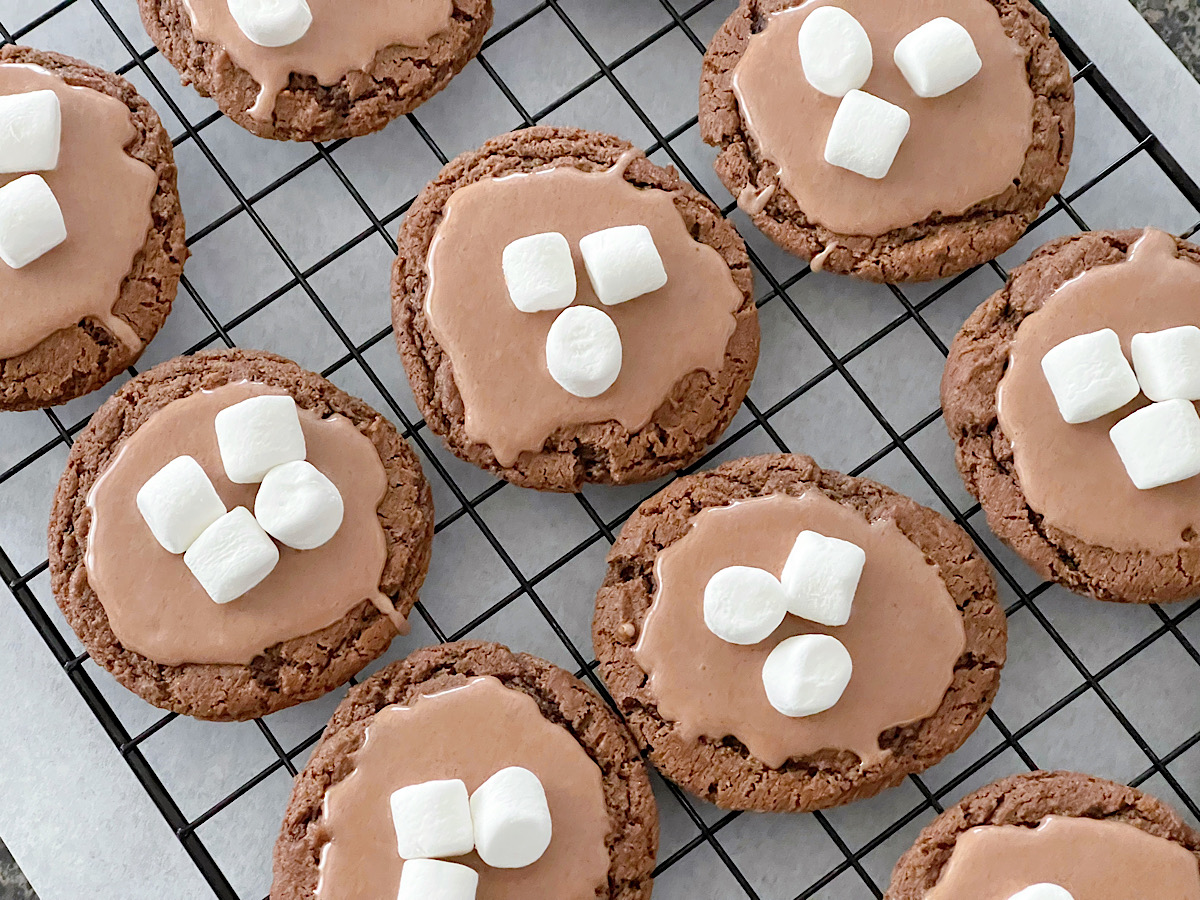 Hot chocolate cookies with hot chocolate glaze, topped with mini marshmallows on a cooling rack.