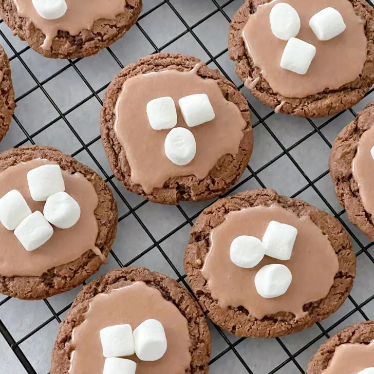 Hot chocolate cookies with hot chocolate glaze, topped with mini marshmallows on a cooling rack.