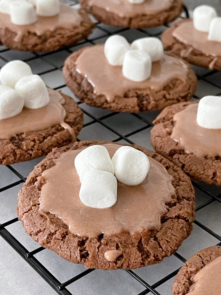 Hot chocolate cookies with hot chocolate glaze, topped with mini marshmallows on a cooling rack.