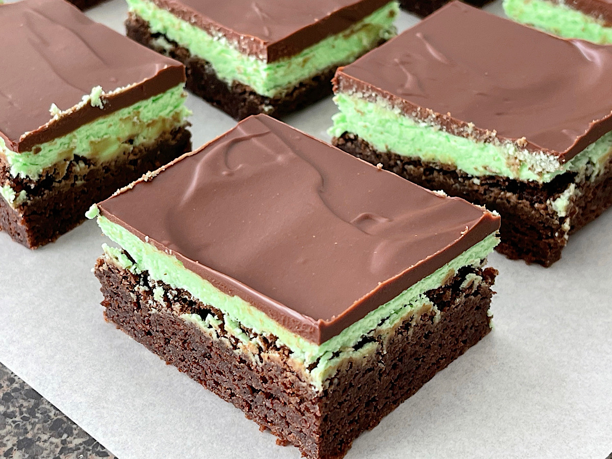 Brownies topped with mint frosting and chocolate ganache on a piece of parchment paper.