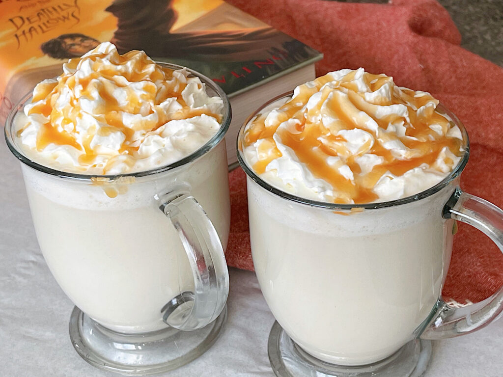 Two mugs of hot butterbeer topped with whipped cream and caramel.