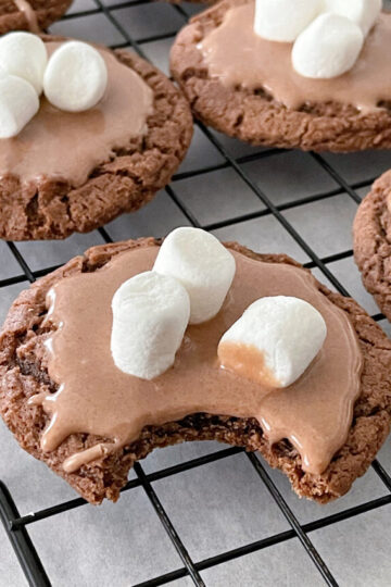 Hot chocolate cookies with hot chocolate glaze, topped with mini marshmallows on a cooling rack.