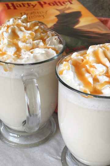 Two mugs of hot butterbeer topped with whipped cream and caramel.
