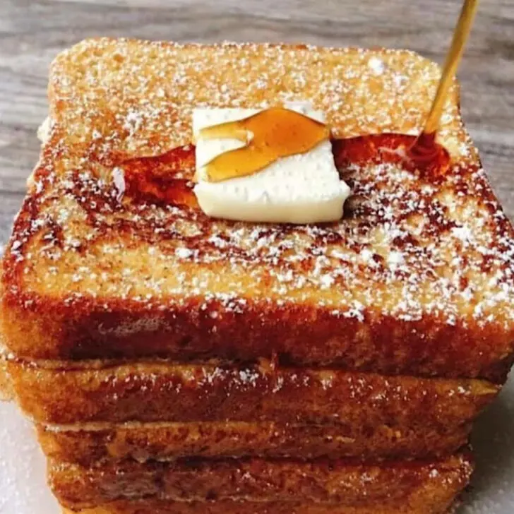 Slices of French Toast stacked on a plate and topped with butter and syrup.