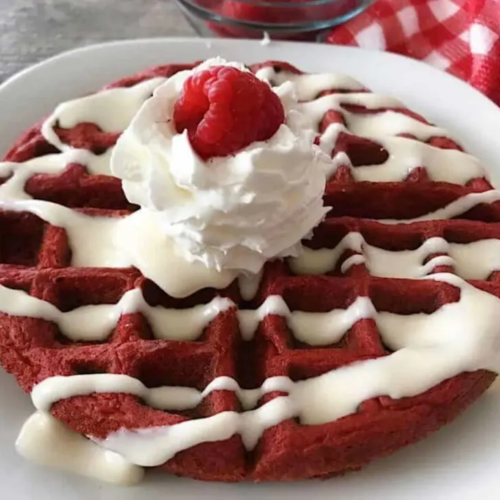 A red velvet cake mix waffle topped with cream cheese syrup, whipped cream, and a raspberry.