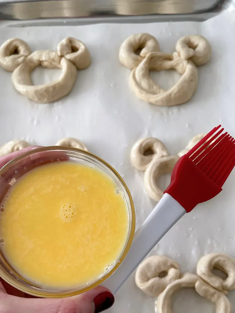 A bowl of beaten egg and a pastry brush with unbaked Mickey pretzels.