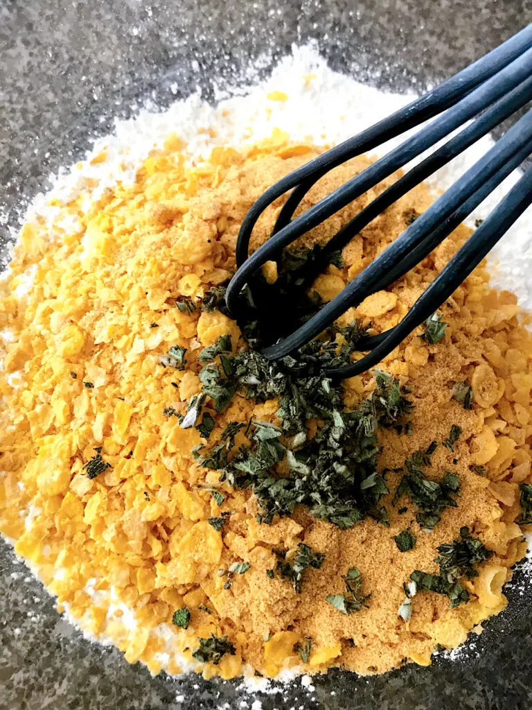 A bowl of crushed corn flakes, fresh sage, flour, and spices for sage fried chicken.