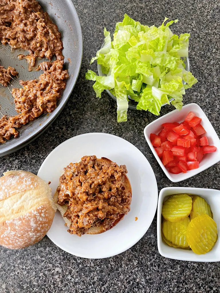 A cheeseburger sloppy joe with a bowl of lettuce, tomatoes, and pickles.
