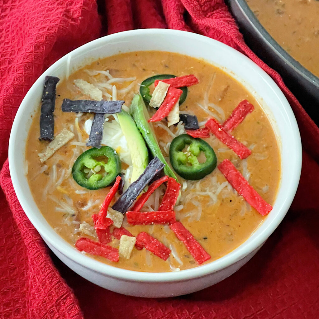 A bowl of cheesy enchilada chili topped with sour cream, jalapenos, and tortilla strips.