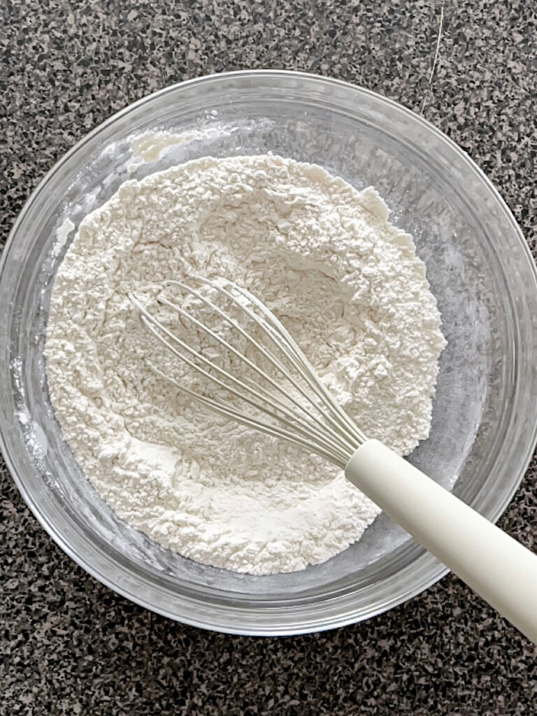 Flour, baking powder, baking soda, and salt mixed together in a bowl with a white whisk.