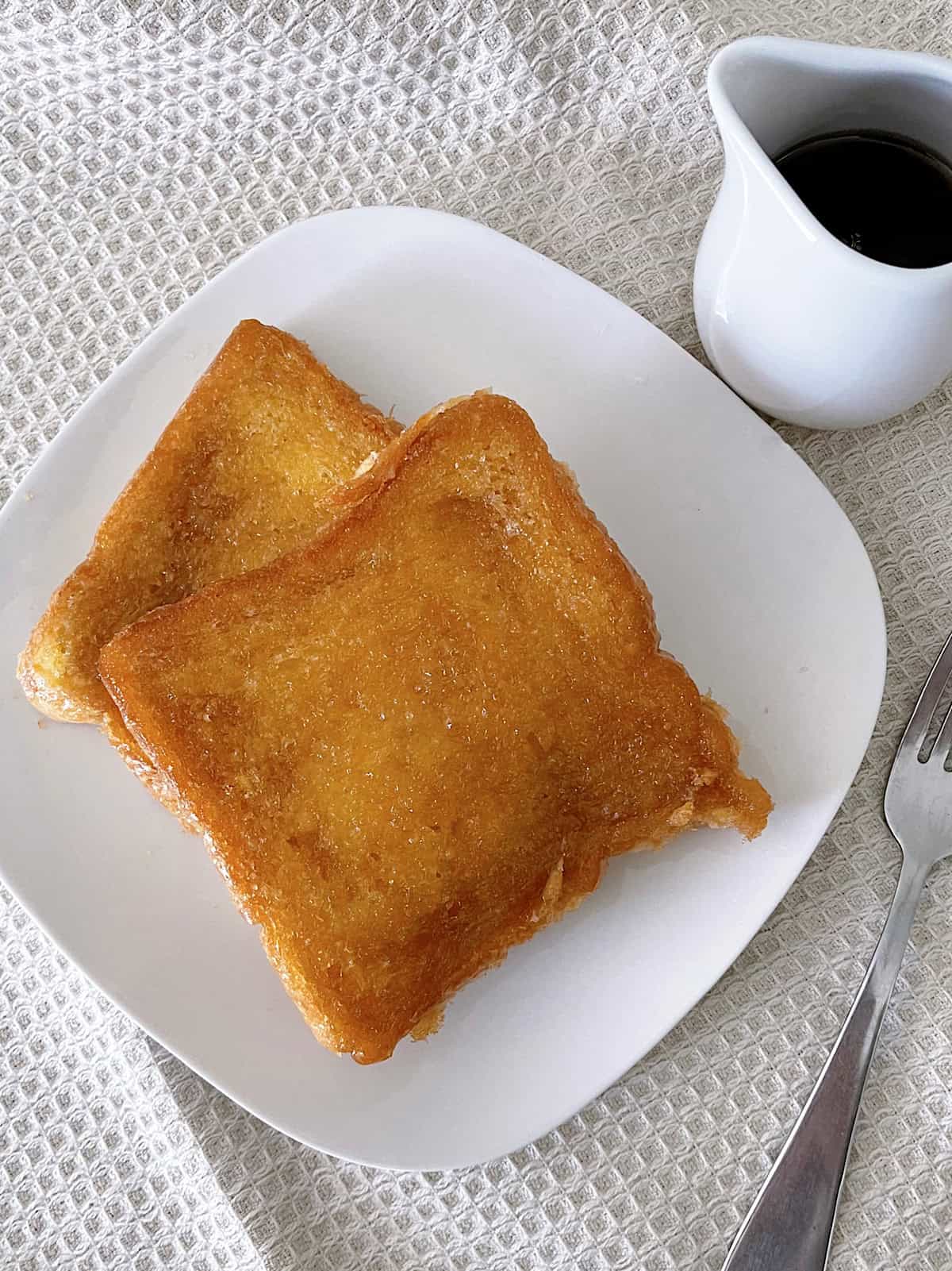 Two slices of Crème Brûlée French Toast on a white plate with a side of maple syrup.