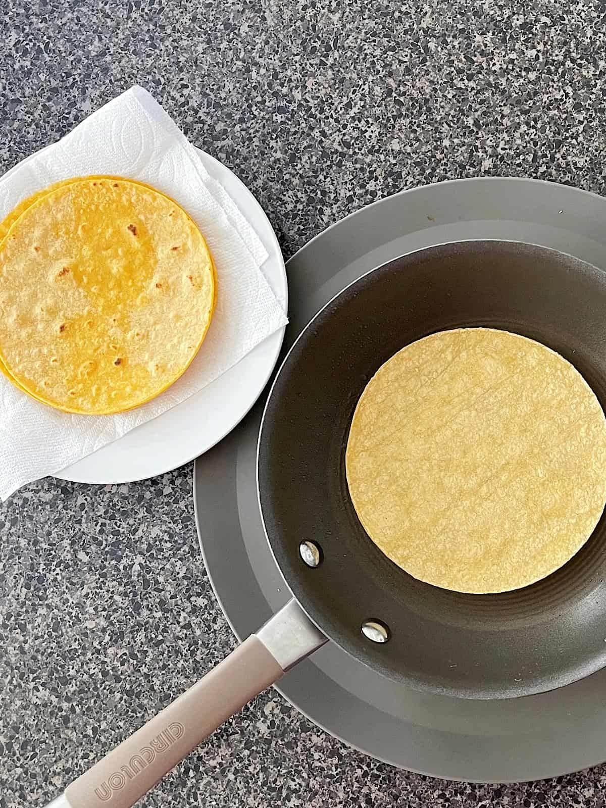 Corn tortillas on a white plate and a tortilla in a small frying pan.