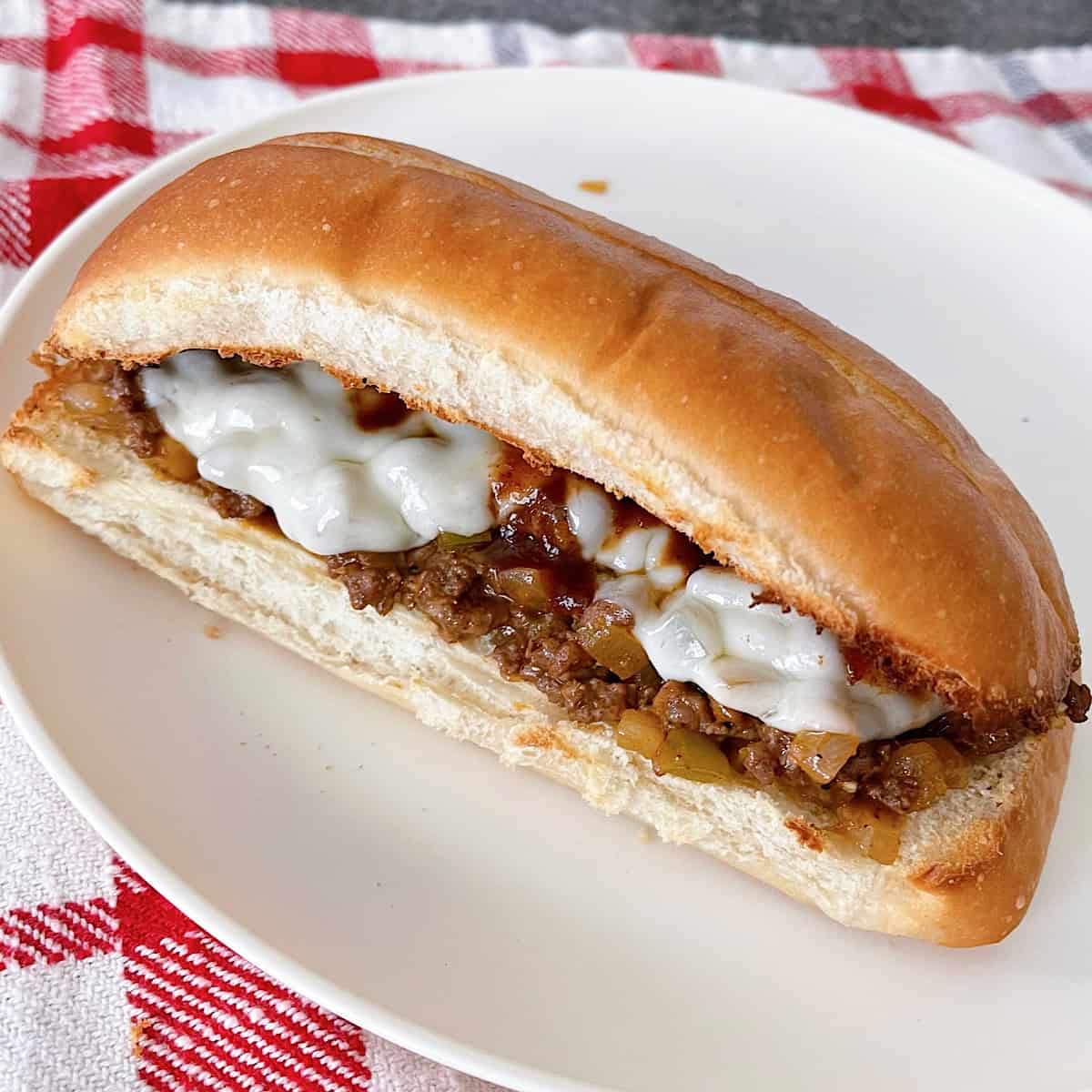 A Philly Cheesesteak Sloppy Joe on a white plate with a red and white kitchen towel.