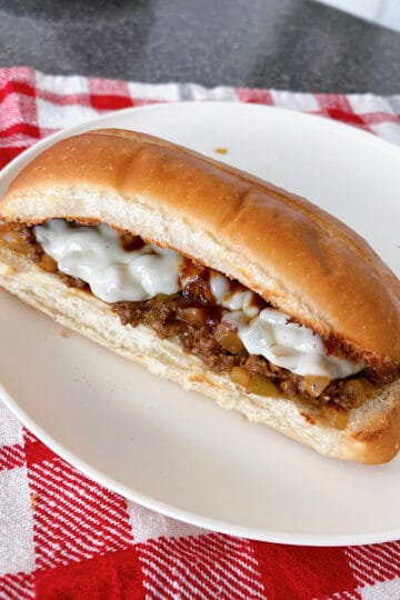 A Philly Cheesesteak Sloppy Joe on a white plate with a red and white kitchen towel.