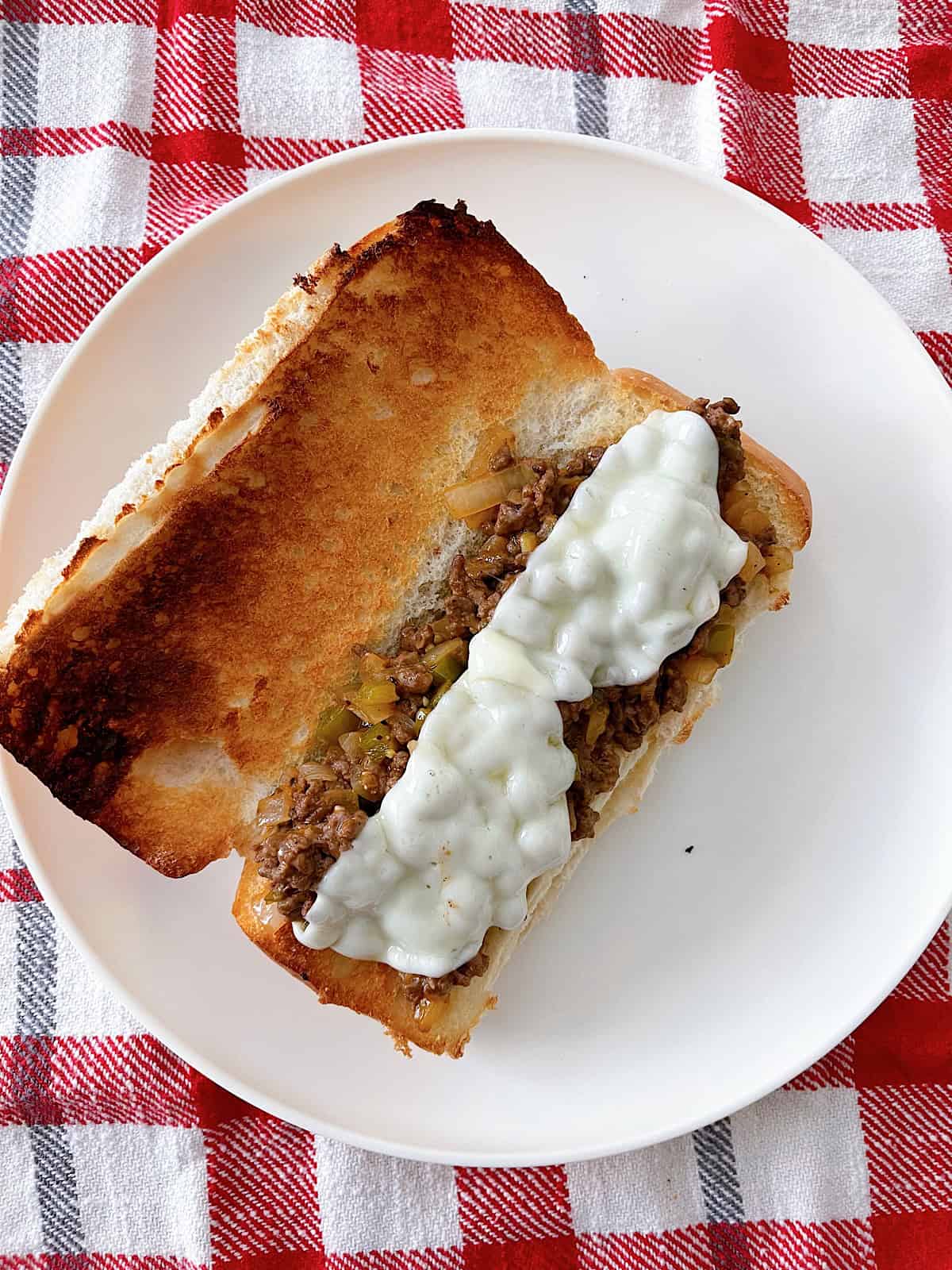 A Philly Cheesesteak Sloppy Joe on a white plate with a red and white kitchen towel.