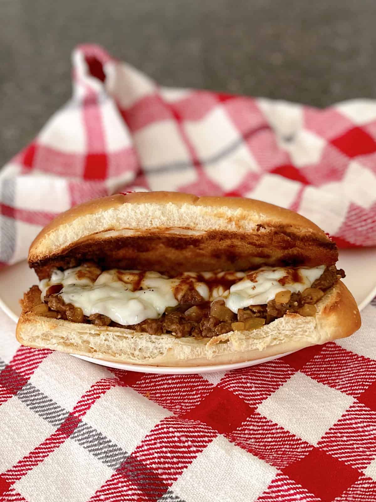 A Philly Cheesesteak Sloppy Joe on a white plate with a red and white kitchen towel.